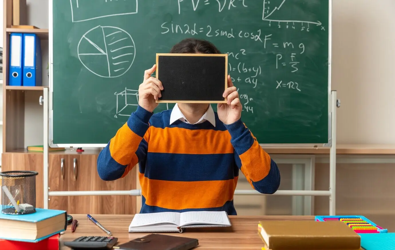 Eine junge kaukasische Geometrielehrkraft mit Brille sitzt im Klassenzimmer an einem Schreibtisch mit Schulutensilien und hält eine Mini-Tafel vor das Gesicht.