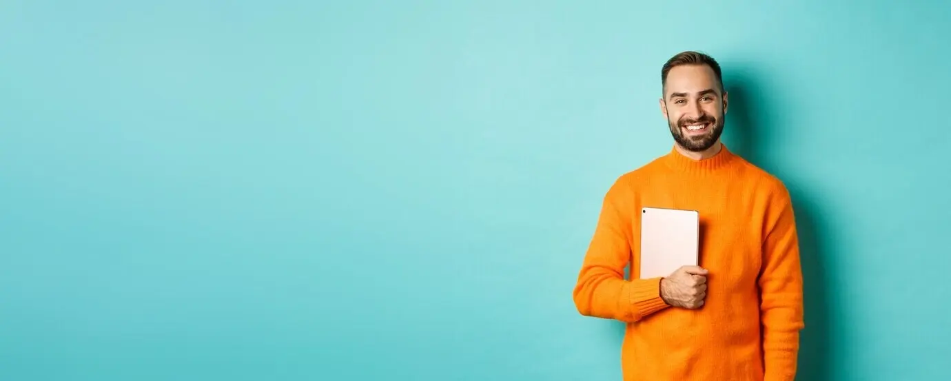 Ein gutaussehender freiberuflicher Mann hält einen Laptop und lächelt, während er glücklich vor hellblauem Hintergrund steht.
